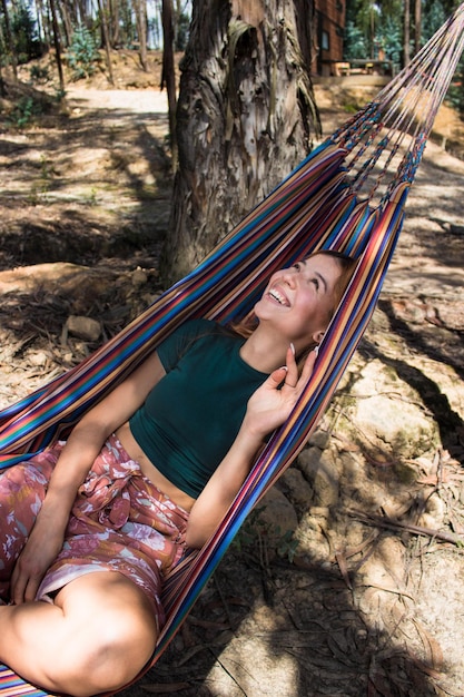 Positive and smiling woman lying in a colorful hammock and posing in the homeyard on a sunny day