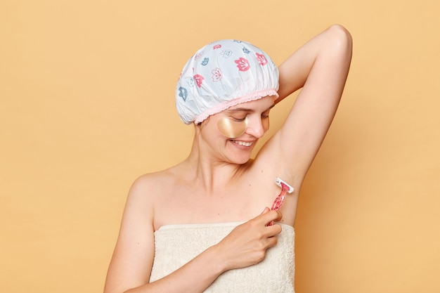 Positive smiling woman wearing shower cap taking care of her skin smiling happily shaving armpit standing isolated over beige background