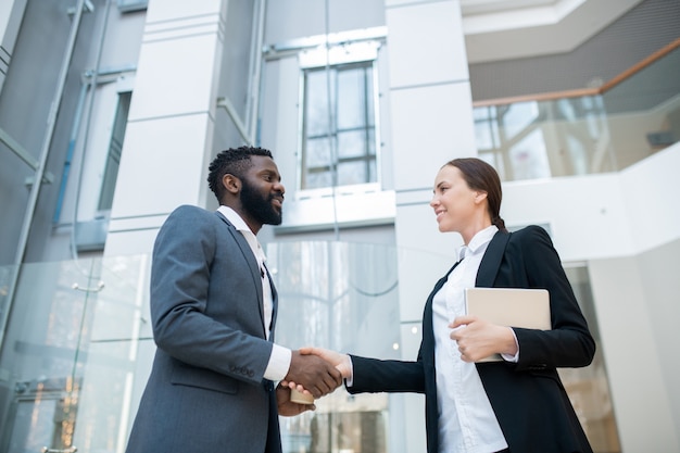 Positive successful Afro-American businessman in suit handshaking with pretty business colleague while greeting her in company