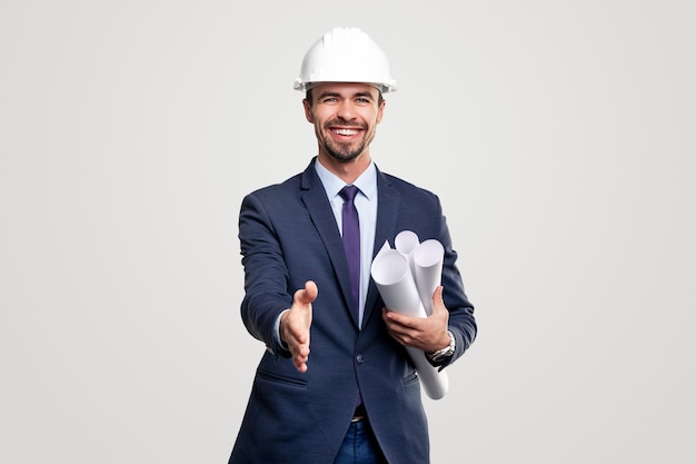 Positive successful male construction engineer in formal suit and hardhat with rolled blueprints outstretching hand for handshake to camera