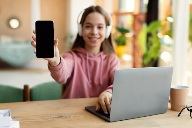 Positive teenage lady demonstrating blank smartphone while studying at desk with laptop at home