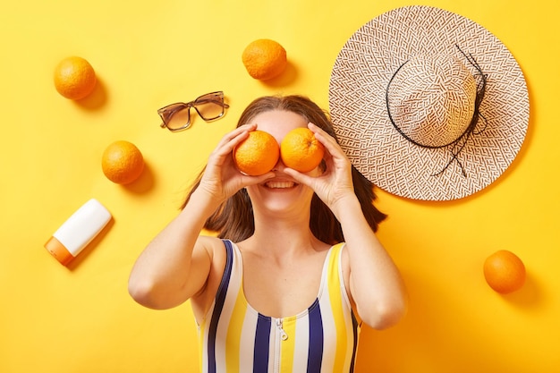 Positive unknown woman wearing striped swimming suit lying with oranges sunscreen on yellow background covering eyes with fruit having fun on her vacation