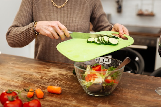 Positive while cooking. Hard-working accurate woman adding chopped cucumbers into glass bowl while finishing her cooking