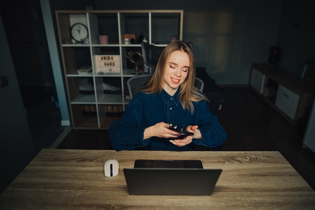 Positive woman in a blue shirt sits at home at her desk with a laptop and uses a smartphone