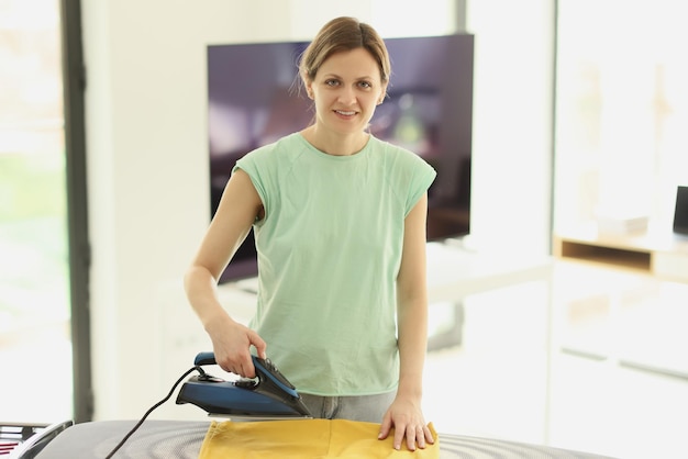 Positive woman irons washed dried clothes with appliance on board at home maid does routine