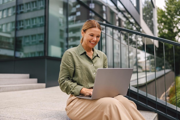 Positive woman manager sitting on stairs with laptop while working on project remotely