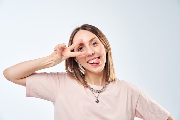 Positive woman portrait. Cheerful happy young woman smiling and show victory gesture on a white wall