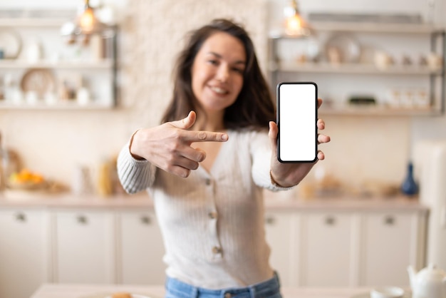 Positive woman showing blank smartphone screen and pointing on it while standing in kitchen