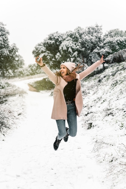 Photo positive woman in warm clothes jumping and taking self portrait on smartphone while having fun in snowy winter woods