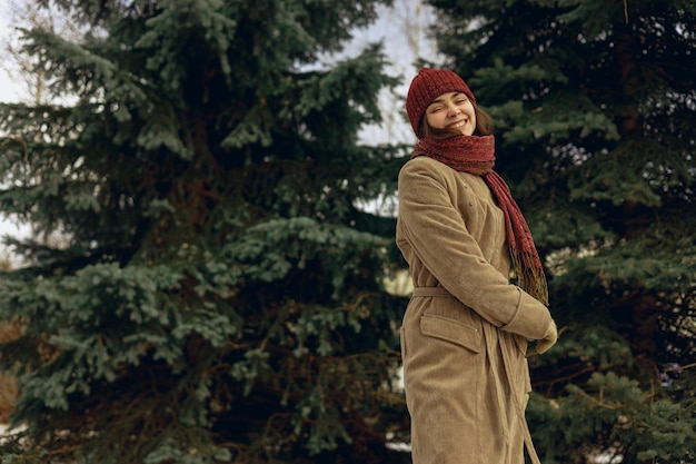 Positive woman wearing warm coat and hat with scarf dancing and having fun in winter park