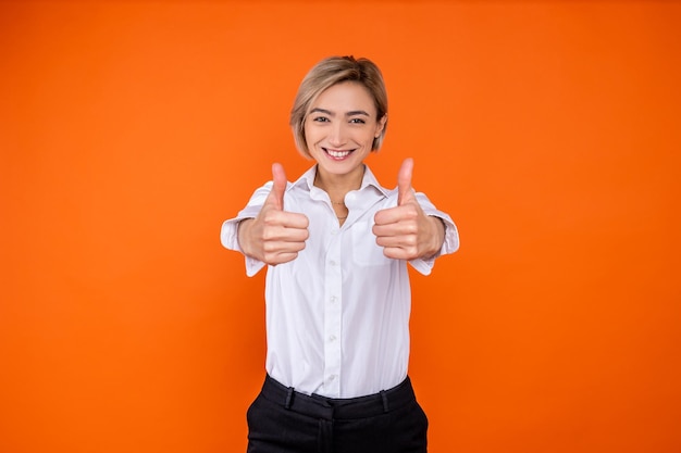 Positive woman wearing white official style shirt showing like gesture and smiling to camera isolated over orange background