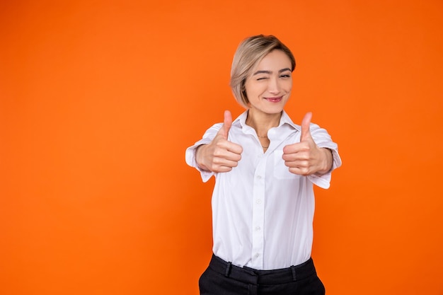 Positive woman wearing white official style shirt showing like gesture and winking to camera isolated over orange background