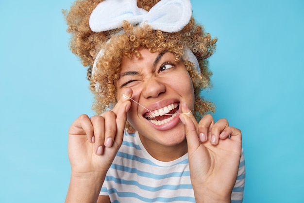 Positive woman with curly blonde hair wears headband and casual striped t shirt flossses teeth uses dental floss winks eye isolated over blue background Oral hygiene and health care concept