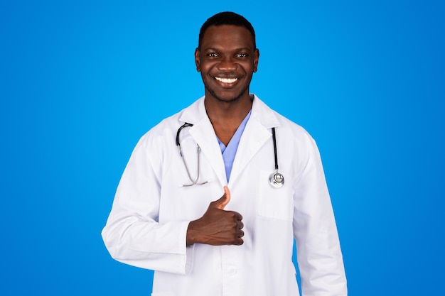 Positive young african american guy doctor in white coat show thumb up, approving ad and offer, isolated on blue studio background. Good health care service, medicine, treatment in clinic