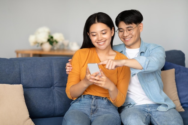 Positive young asian couple sitting on couch using smartphone