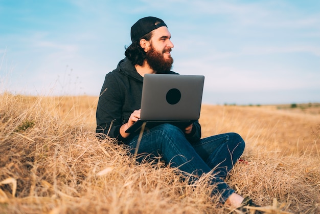 Positive young bearded  man working on the computer on a field