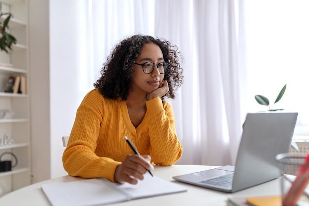 Positive young black female student having online lesson on laptop computer writing down information