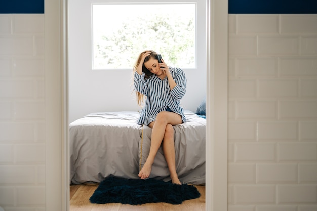 positive young female siting in bed with cozy clothes and having a phone conversation