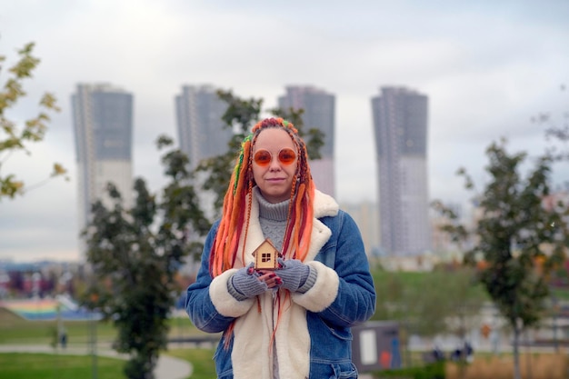 Positive young female in warm clothes and with long dyed dreadlocks holding small wooden house and looking at camera against blurred skyscrapers in city