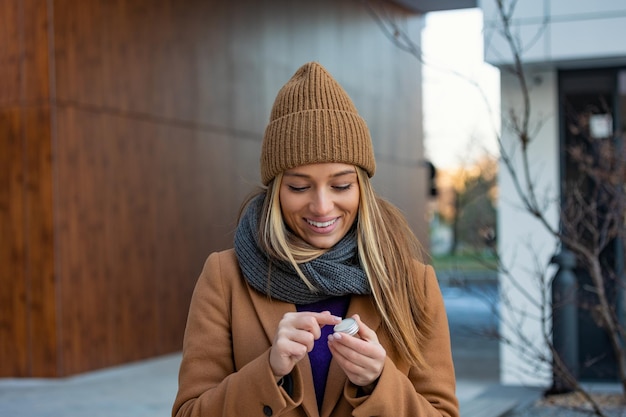 Positive young female with long blond hair in warm coat applying lip balm while standing on street near building during stroll