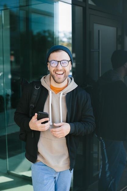 Positive young guy holding smartphone in his hands looking at camera standing on street Bearded man in eyeglass wears casual clothes Cell phone usage concept