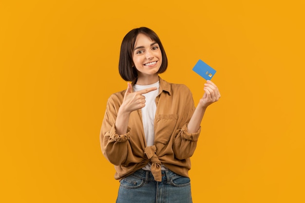 Positive young lady pointing at credit card recommending banking service posing over yellow background and smiling