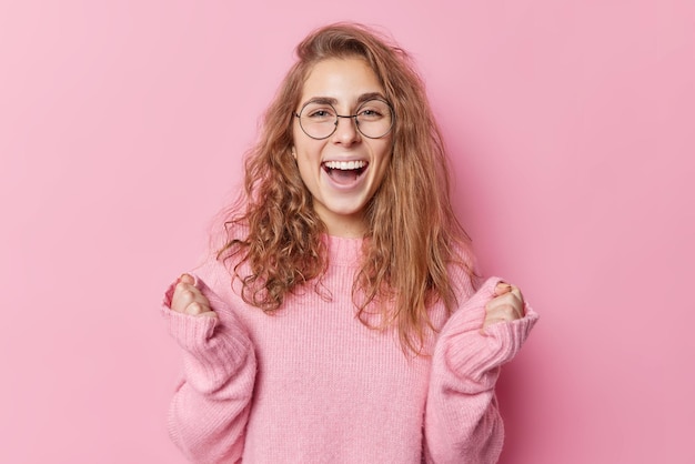 Positive young long haired woman with pleasant appearance clenches fists exclaims from joy wears round spectacles and casual loose jumper celebrates personal achievements isolated on pink background