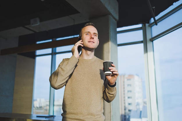 Positive young man smiling and talking on mobile phone and drinking coffee while resting while standing in office Lunch break during work