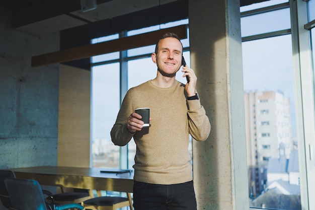 Positive young man smiling and talking on mobile phone and drinking coffee while resting while standing in office Lunch break during work