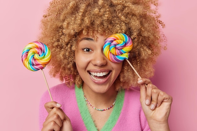 Positive young woman covers eye with multicolored caramel lollipop enjoys eating sweet food smiles gladfully dressed in casual wear isolated over pink background cant wait to get bite of candy