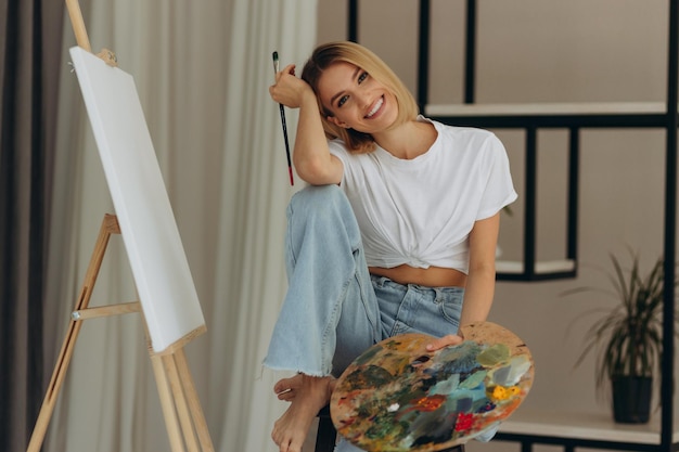 Positive young woman holding paintbrush and palette with colorful paints near easel Artist