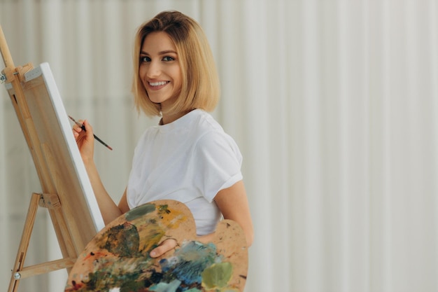 Positive young woman holding paintbrush and palette with colorful paints near easel Artist