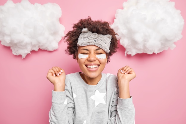 Positive young woman rejoices good news smiles broadly keeps eyes closed clenches hands in fists wears pajama and sleepmask isolated over pink wall