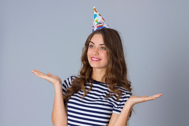 Positive young woman wearing in stripped T-shirt and celebration cap on grey background in studio