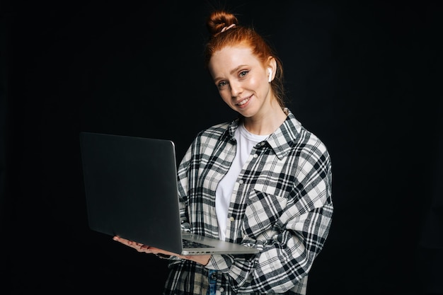 Positive young woman wearing wireless earphones holding laptop computer and looking at camera