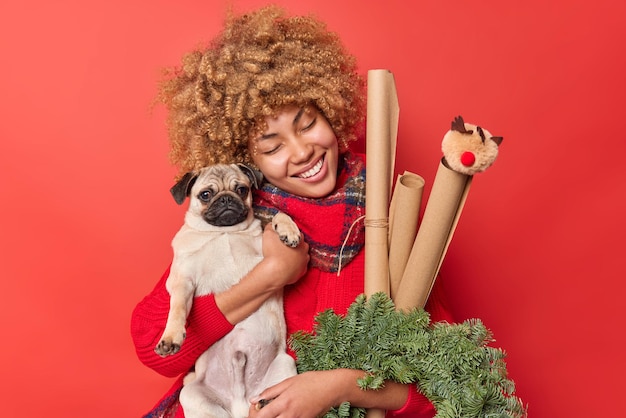 Photo positive young woman with blonde curly hair embraces with love her favorite pug dog carries green spruce wreath and paper rollers keeps eyes closed smiles gently isolated over red background