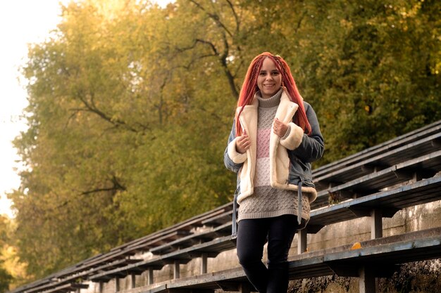 Positive young woman with dreadlocks looking at camera smiling posing near old shabby bench of street bleacher in golden autumn