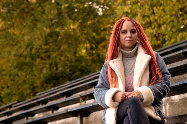 Positive young woman with dreadlocks looking at camera smiling sitting on old shabby bench of street bleacher in golden autumn