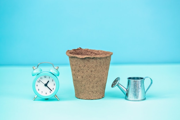 A pot for seedlings on a blue background with an alarm clock and a watering can.