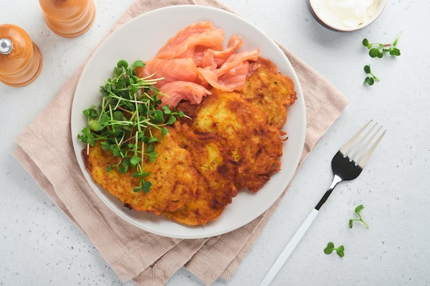 Potato pancakes Fried homemade potato pancakes or latkes with cream green onions microgreens red salmon and sauce in rustic plate on white table background Rustic style Healthy food Top view