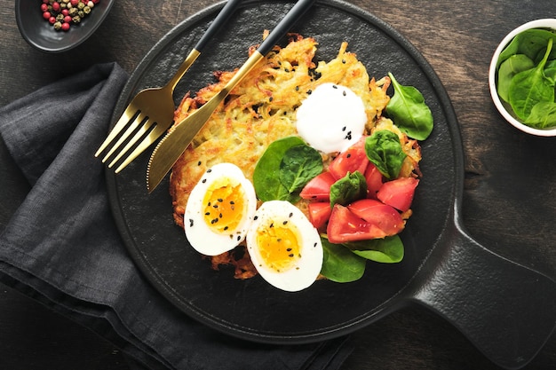 Potatoes latkes with sour cream spinach salad tomatoes and boiled eggs on dark wooden old table background Homemade tasty potato pancakes Delicious food for breakfast Top view