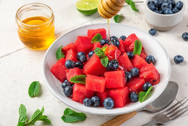 Pouring honey in watermelon salad with blueberries and mint in a plate on white table Healthy summer dessert