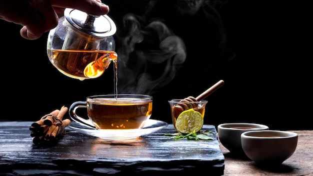 Pouring hot herbal tea into glass teacup with various herbs on black stone slate in dark background