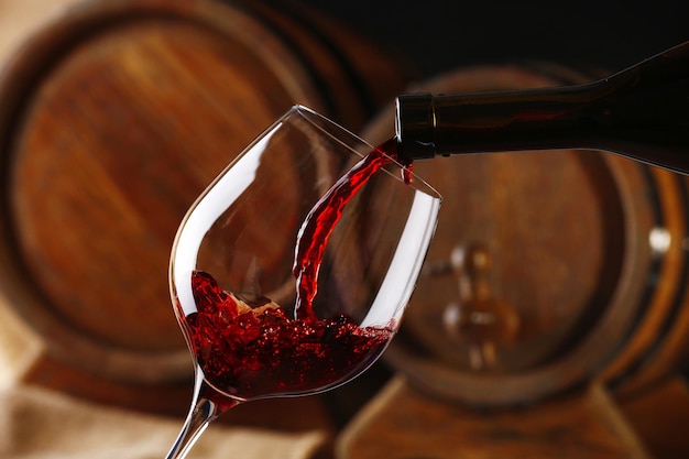 Pouring red wine from bottle into glass with wooden wine casks on background