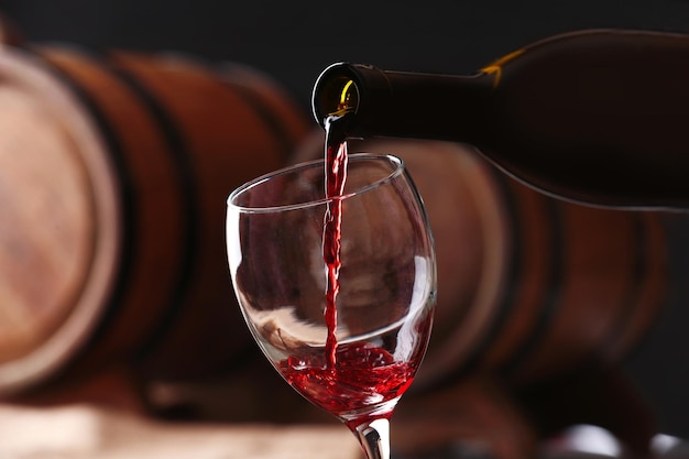 Pouring red wine from bottle into glass with wooden wine casks on background