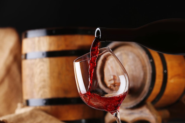 Pouring red wine from bottle into glass with wooden wine casks on background