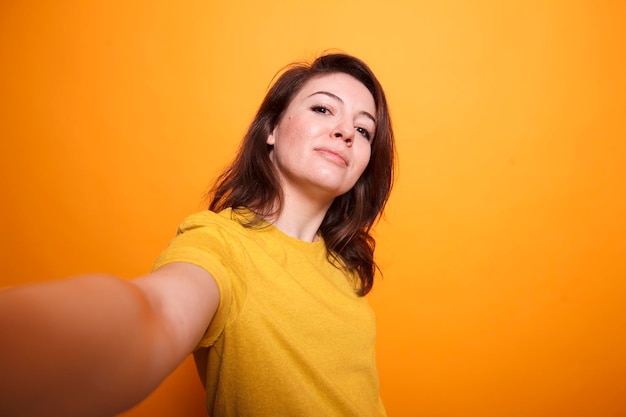 Photo pov of caucasian female influencer wearing yellow tshirt taking selfie with camera while on isolated orange background brunette carefree vlogger smiling at camera while recording a daily vlog