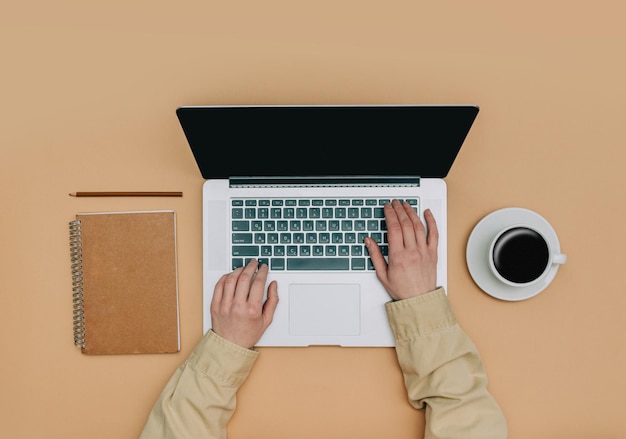 POV view on female hands over laptop computer next to cup of coffee and notebook on brown background