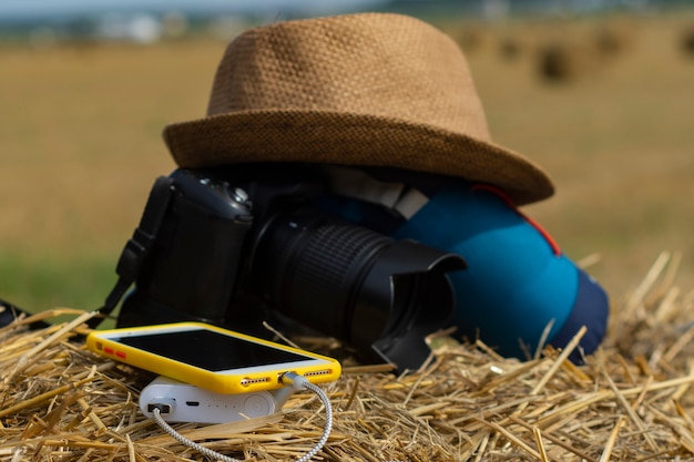 Photo power bank charges a smartphone on the background of a camera with a bag and a hat on a background of hay in nature.