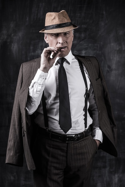 Powerful boss. Serious senior man in hat and suspenders smoking cigar and looking at you while standing against dark background